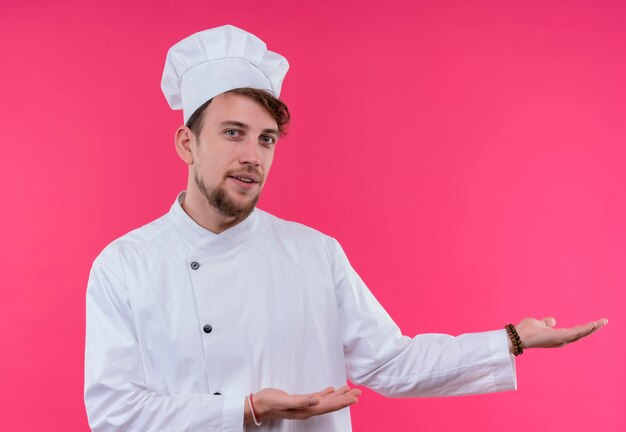 Un joven chef barbudo positivo en uniforme blanco que presenta e invita a venir con la mano mientras mira en una pared rosa