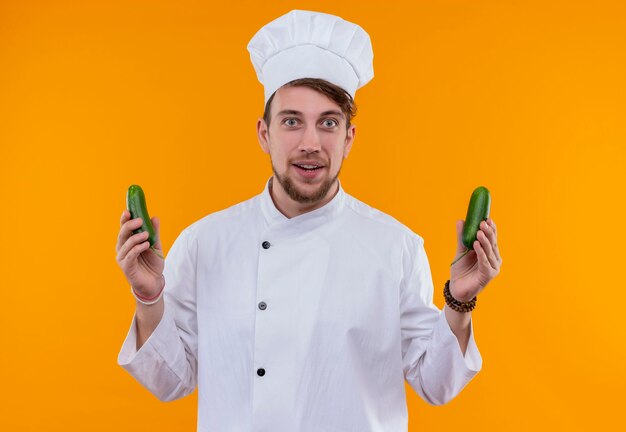 Un joven chef barbudo positivo en uniforme blanco sosteniendo pepinos frescos verdes mientras mira en una pared naranja