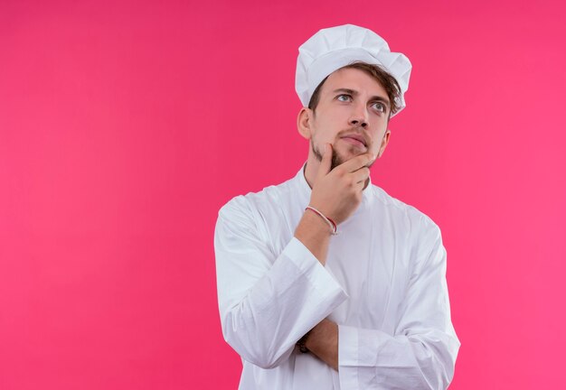 Un joven chef barbudo serio en uniforme blanco pensando mientras sostiene la mano en la barbilla en una pared rosa