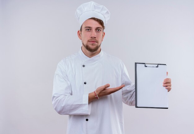 Un joven chef barbudo serio en uniforme blanco que muestra una carpeta en blanco mientras mira en una pared blanca