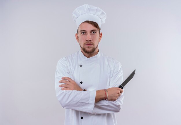 Un joven chef barbudo serio en uniforme blanco sosteniendo un cuchillo mientras mira en una pared blanca