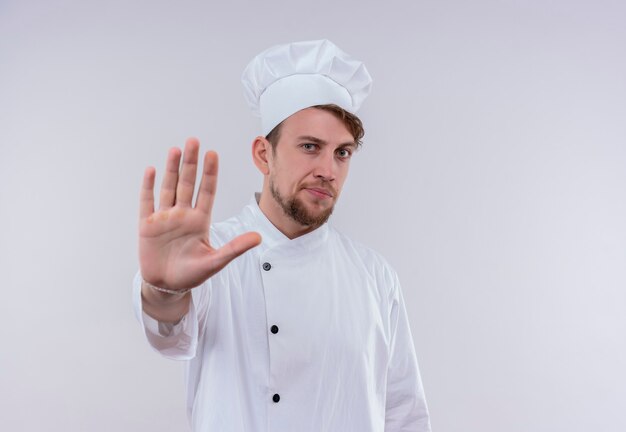 Un joven chef barbudo serio vestido con uniforme de cocina blanco y sombrero mostrando gesto de parada con la mano mientras mira en una pared blanca