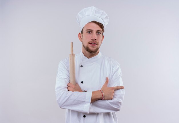 Un joven chef barbudo serio vestido con uniforme de cocina blanco y sombrero sosteniendo un rodillo y apuntando hacia un lado mientras mira en una pared blanca