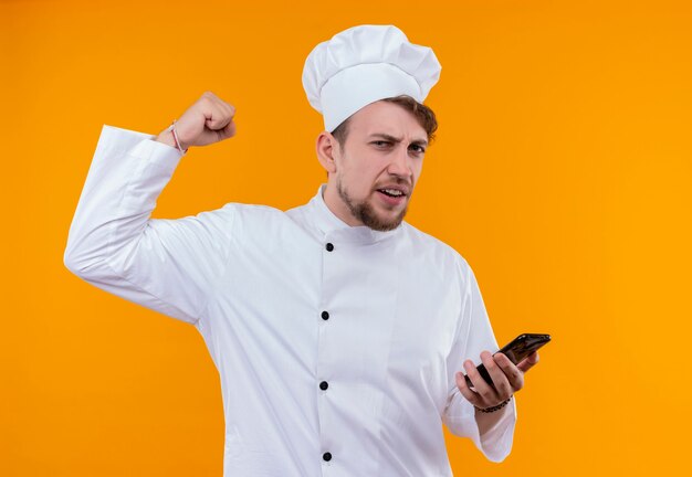 Un joven chef barbudo con uniforme blanco levantando el puño cerrado y sosteniendo el teléfono móvil mientras mira en una pared naranja