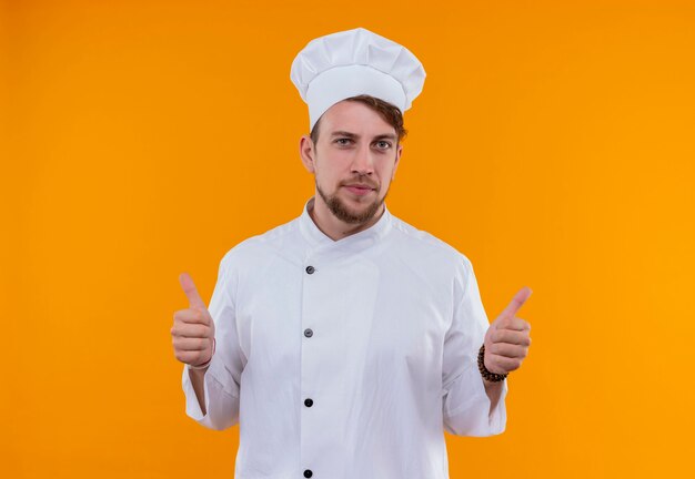 Un joven chef barbudo con uniforme blanco mostrando los pulgares hacia arriba mientras mira en una pared naranja