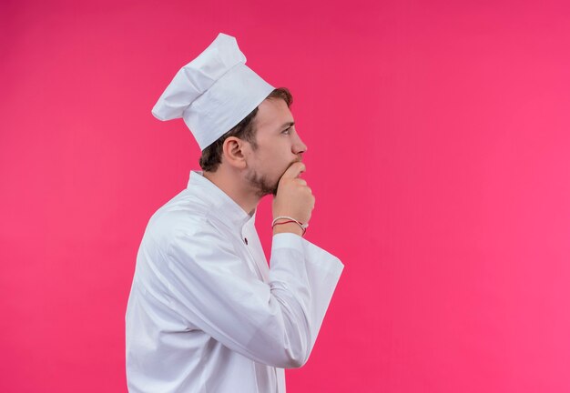Un joven chef barbudo con uniforme blanco pensando con la mano en la barbilla y mirando de lado mientras está de pie sobre una pared rosa