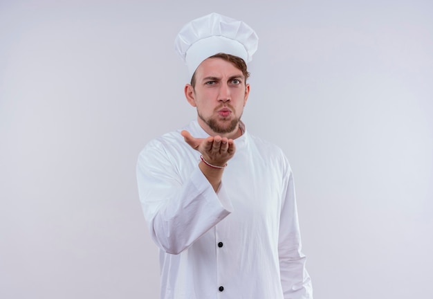 Un joven chef barbudo vestido con uniforme de cocina blanco y sombrero enviando un beso mientras está de pie y mirando una pared blanca