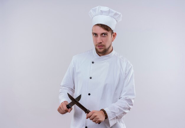 Un joven chef barbudo vestido con uniforme de cocina blanco y sombrero mostrando el signo x con un cuchillo mientras mira en una pared blanca