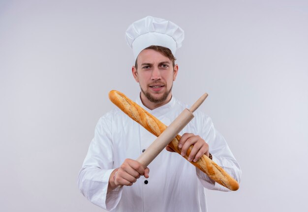 Un joven chef barbudo vestido con uniforme de cocina blanco y sombrero sosteniendo pan baguette con rodillo en signo x mientras mira en una pared blanca