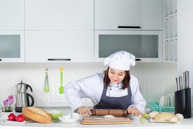 Joven chef de comis femenina ocupada en uniforme de pie detrás de la mesa preparando pasteles en la cocina blanca