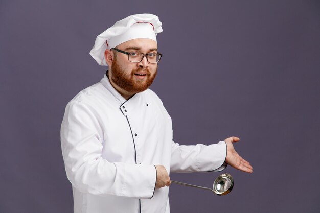 Joven chef complacido con uniforme de anteojos y gorra de pie en la vista de perfil mirando a la cámara apuntando con la mano y con un cucharón a un lado aislado en un fondo morado