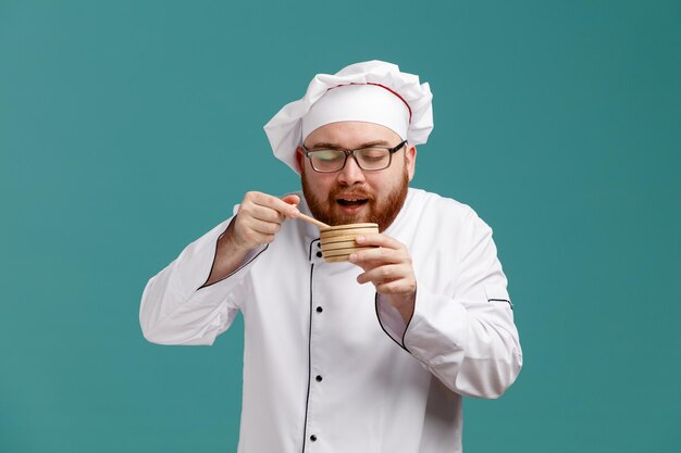 Un joven chef complacido con uniforme de anteojos y gorra sosteniendo un tazón de madera con un cucharón de miel cerca de la boca agarrando una cuchara de miel que huele el aroma de la miel con los ojos cerrados