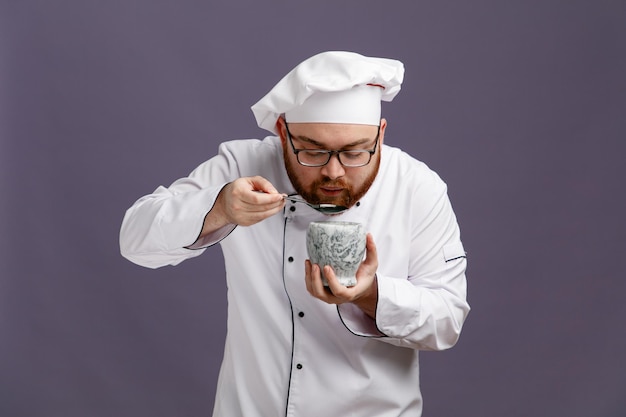 Joven chef concentrado con uniforme de anteojos y gorra sosteniendo un tazón que finge comer del tazón con una cuchara mirando hacia abajo aislado en un fondo morado