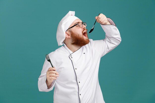 Un joven chef concentrado con uniforme de anteojos y gorra sosteniendo un tenedor y una cuchara cerca de la boca finge comer algo aislado de fondo azul