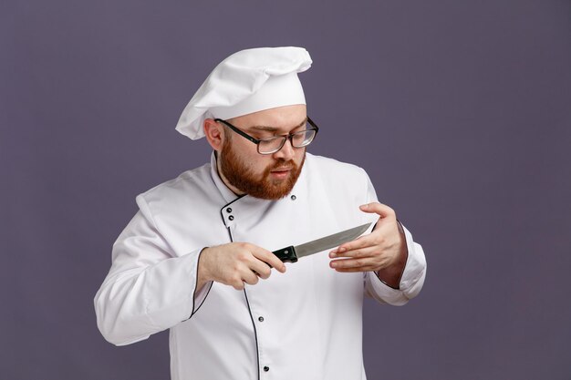 Joven chef concentrado con uniforme de gafas y gorra sosteniendo y mirando el cuchillo cortándose la mano con un cuchillo aislado en un fondo morado