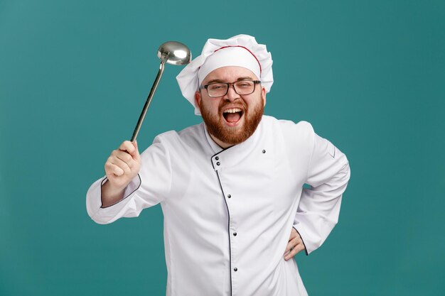 Un joven chef confiado con uniforme de anteojos y gorra que muestra un cucharón mirando a la cámara mientras mantiene la mano en la cintura aislada en el fondo azul