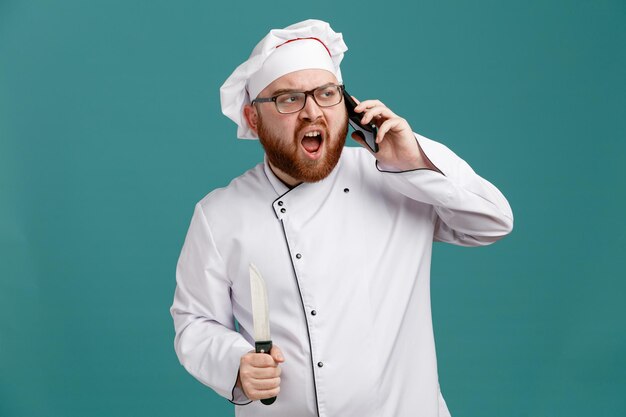 Un joven chef descontento con uniforme de gafas y gorra sosteniendo un cuchillo mirando al lado hablando por teléfono aislado de fondo azul