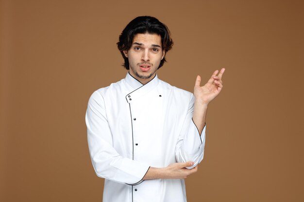 Un joven chef despistado que vestía uniforme manteniendo la mano bajo el codo apuntando hacia un lado aislado de fondo marrón