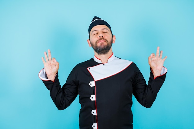 El joven chef está meditando cerrando los ojos y levantando las manos sobre fondo azul.