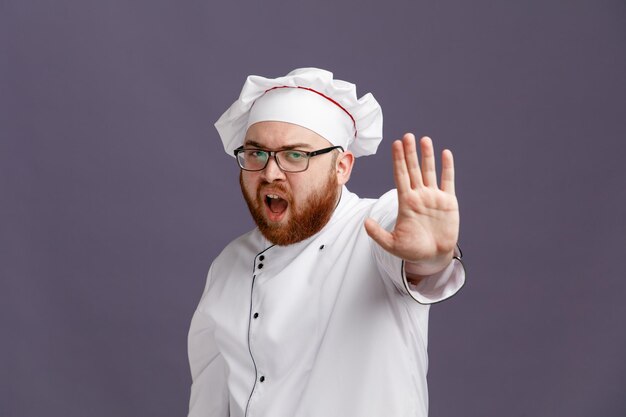Joven chef frunciendo el ceño con uniforme de gafas y gorra mirando a la cámara mostrando un gesto de parada aislado en un fondo morado