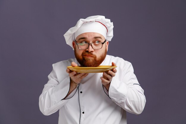Joven chef inseguro con uniforme de anteojos y gorra mirando al costado mientras sostiene el plato debajo de la barbilla aislado en un fondo morado