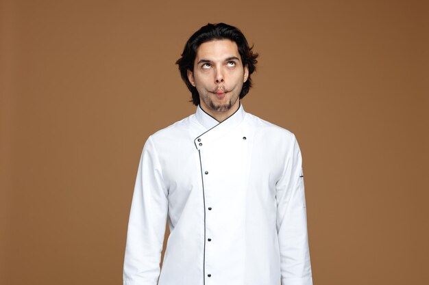 un joven chef loco con uniforme mirando hacia arriba haciendo cara de pez aislado de fondo marrón