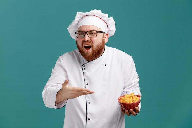 Un joven chef molesto con uniforme de anteojos y gorra sosteniendo un tazón de pasta de macarrones apuntándolo con la mano mirando al lado aislado en el fondo azul