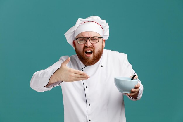 Un joven chef molesto con uniforme de anteojos y gorra sosteniendo un tazón vacío con una cuchara apuntando con la mano al tazón mirando la cámara aislada en el fondo azul