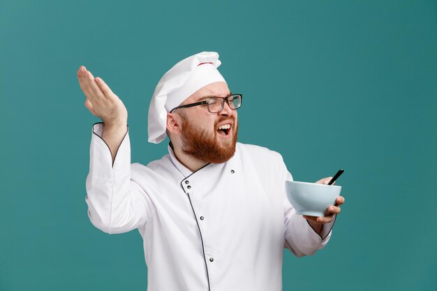 Un joven chef molesto con uniforme de anteojos y gorra sosteniendo un tazón vacío con una cuchara mirando al costado mostrando la mano vacía cerca de la cabeza aislada en el fondo azul