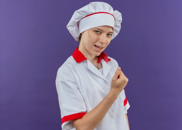 Joven chef mujer rubia alegre en uniforme de chef mantiene el puño en alto aislado en la pared violeta