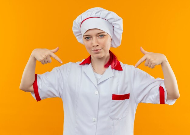 Joven chef mujer rubia complacida en uniforme de chef apunta hacia abajo con ambas manos aisladas en la pared naranja