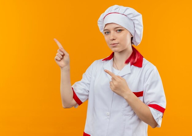 Joven chef mujer rubia complacida en uniforme de chef apunta al lado aislado en la pared naranja