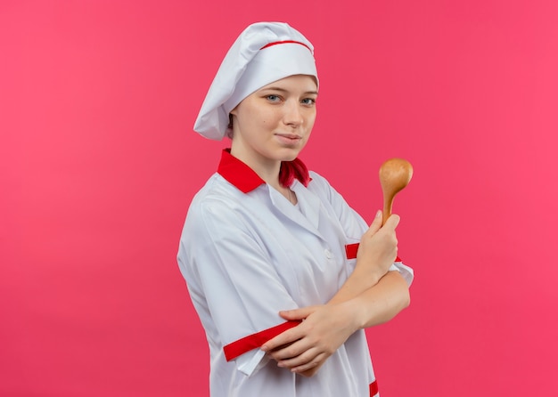 Joven chef mujer rubia complacida en uniforme de chef se para de lado con los brazos cruzados y sostiene una cuchara de madera aislada en la pared rosa