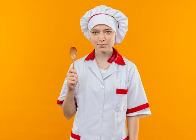 Joven chef mujer rubia complacida en uniforme de chef sostiene una cuchara de madera aislada en la pared naranja