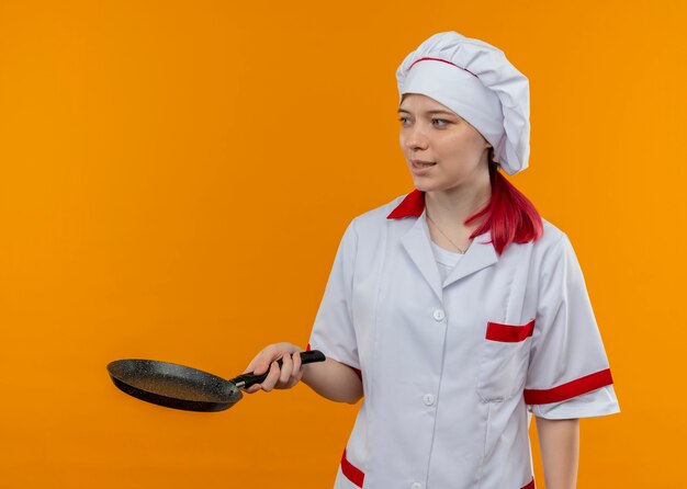 Joven chef mujer rubia complacida en uniforme de chef sostiene una sartén y mira el lado aislado en la pared naranja