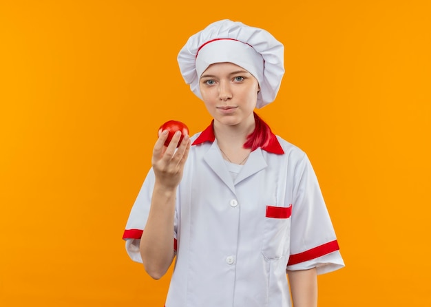 Joven chef mujer rubia complacida en uniforme de chef sostiene tomate y parece aislado en la pared naranja