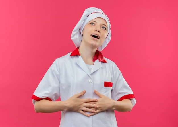 Joven chef mujer rubia complacida en uniforme de chef sostiene el vientre con ambas manos aisladas en la pared rosa