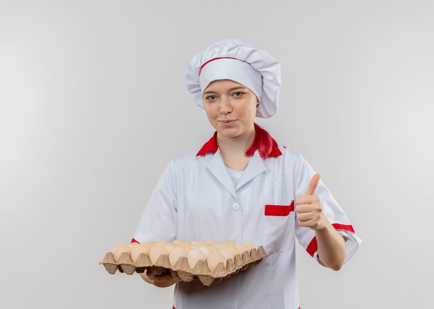 Joven chef mujer rubia complacida en uniforme de chef tiene lote de huevos y pulgares arriba aislados en la pared blanca