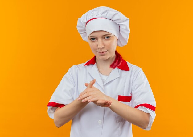 Joven chef mujer rubia complacida en uniforme de chef tiene las manos juntas aisladas en la pared naranja