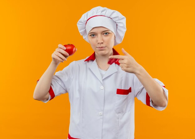 Joven chef mujer rubia confiada en uniforme de chef sostiene y apunta a tomate aislado en pared naranja