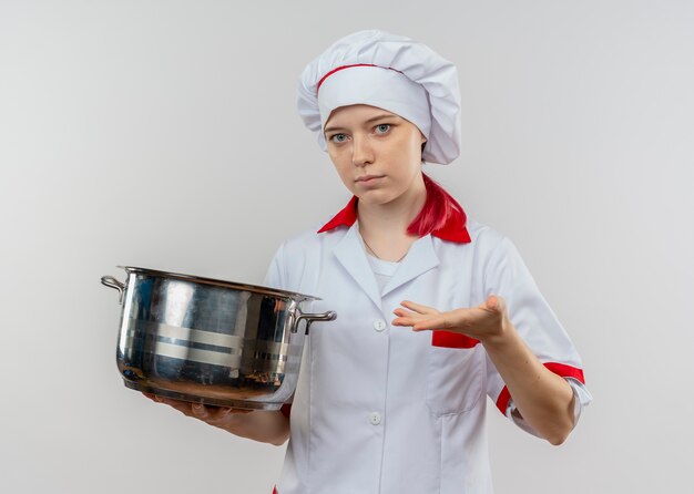 Joven chef mujer rubia confiada en uniforme de chef sostiene olla aislada en la pared blanca