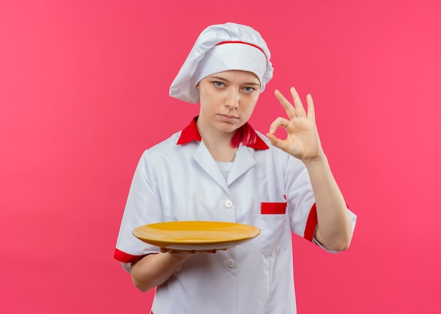 Joven chef mujer rubia confiada en uniforme de chef sostiene placa y gestos ok signo de mano aislado en la pared rosa