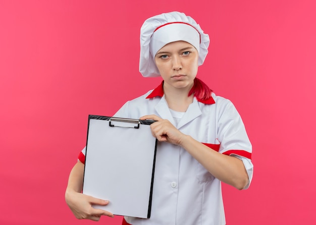 Joven chef mujer rubia confiada en uniforme de chef sostiene el portapapeles aislado en la pared rosa