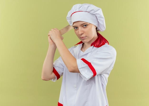 Joven chef mujer rubia confiada en uniforme de chef sostiene el rodillo con ambas manos aisladas en la pared verde