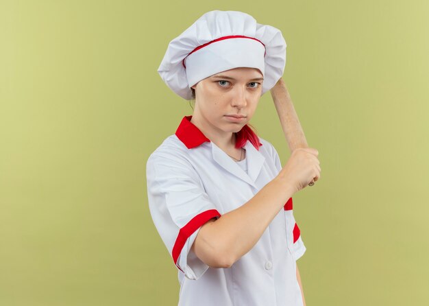 Joven chef mujer rubia confiada en uniforme de chef sostiene el rodillo en la mano y parece aislado en la pared verde