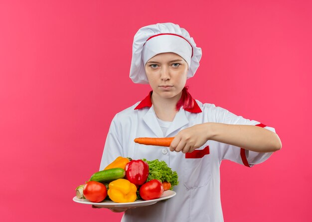 Joven chef mujer rubia confiada en uniforme de chef sostiene verduras en plato y zanahoria en mano aislado en pared rosa