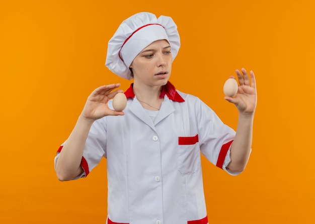 Joven chef mujer rubia confundida en uniforme de chef tiene huevos aislados en la pared naranja