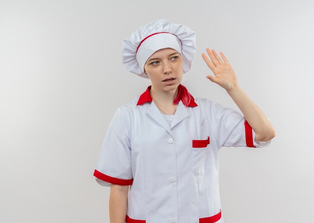 Joven chef mujer rubia molesta en uniforme de chef levanta la mano y mira el lado aislado en la pared blanca