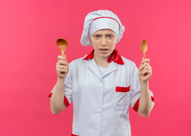 Joven chef mujer rubia molesta en uniforme de chef sostiene cucharas de madera aisladas en la pared rosa