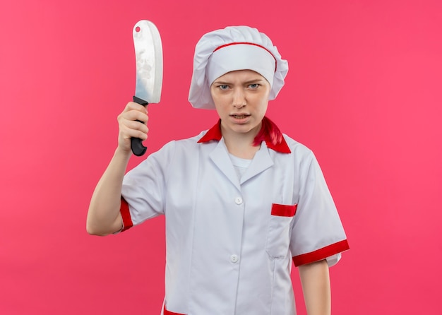 Joven chef mujer rubia molesta en uniforme de chef sostiene un cuchillo aislado en la pared rosa